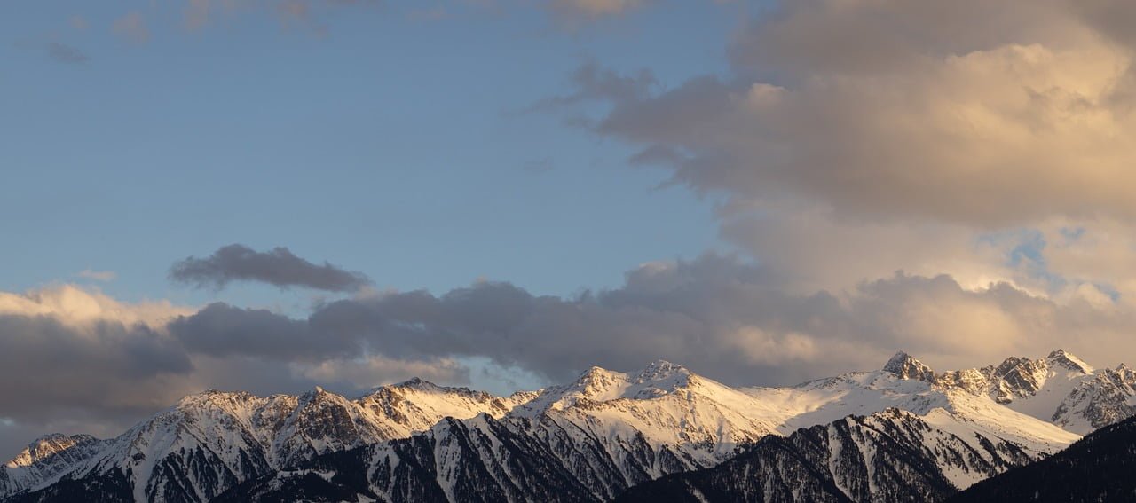 Snow-capped mountain peaks bathed in soft golden light from a setting or rising sun, with scattered clouds in the sky.