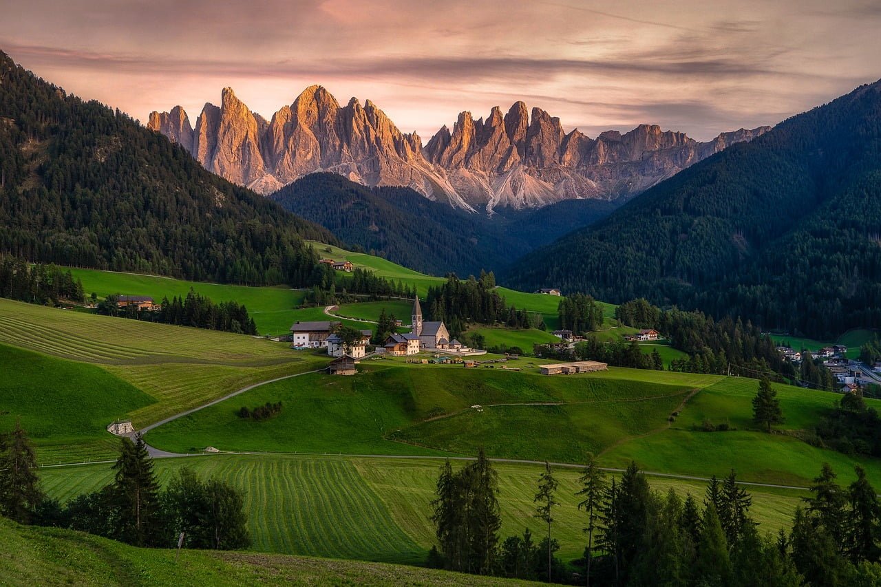 A picturesque landscape with the sun setting on a quaint village nestled amongst lush green fields and towering, jagged mountain peaks in the background.