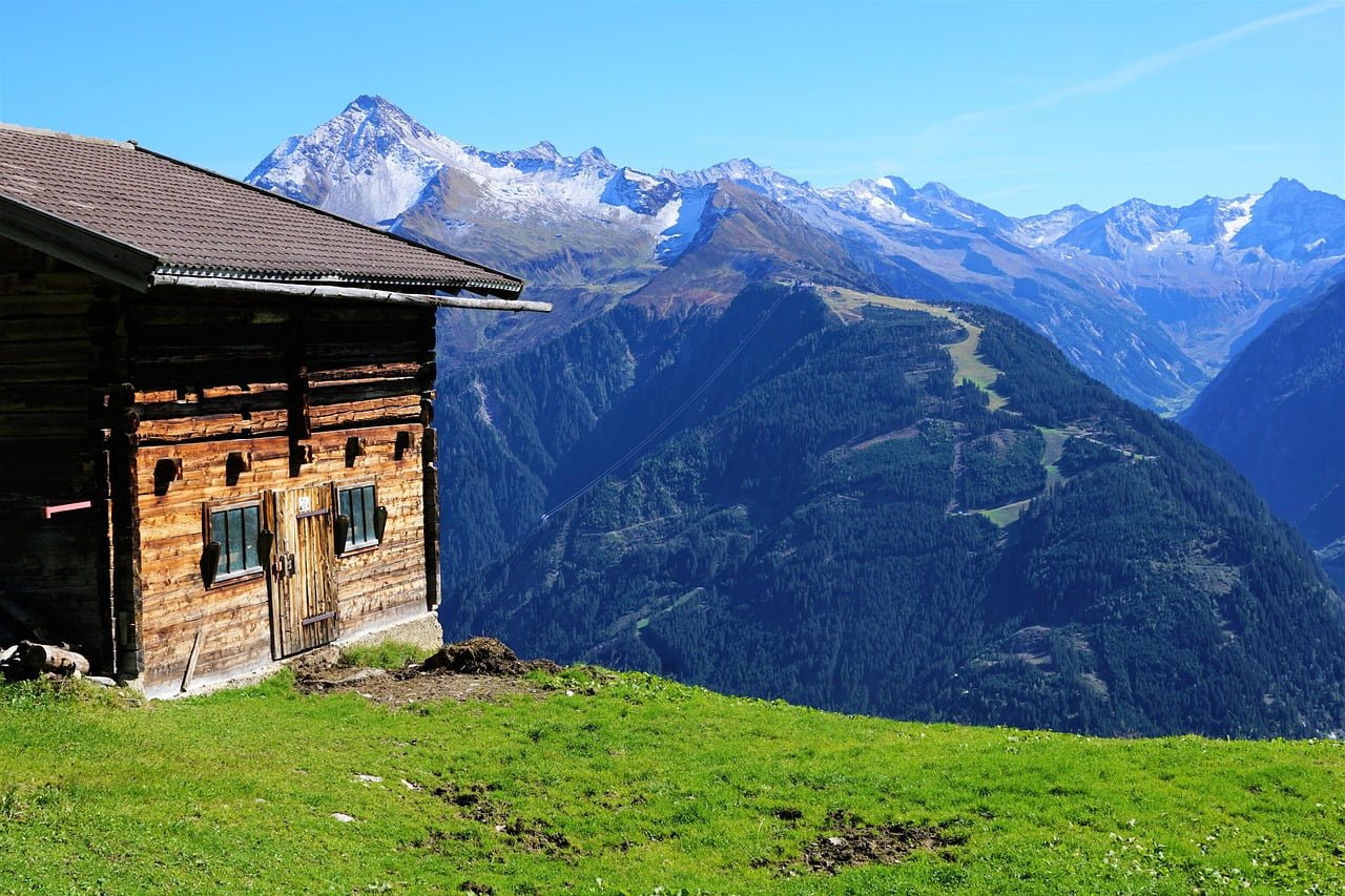 A rustic wooden cabin on a grassy hillside with scenic views of snow-capped mountains in the distance.