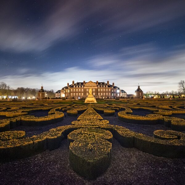 An illuminated baroque palace at night with an ornate formal garden in the foreground and a starry sky with moving clouds above.
