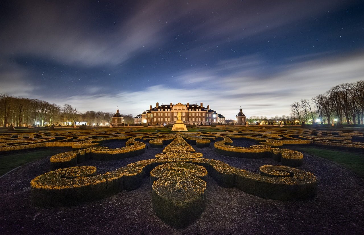 An illuminated baroque palace at night with an ornate formal garden in the foreground and a starry sky with moving clouds above.