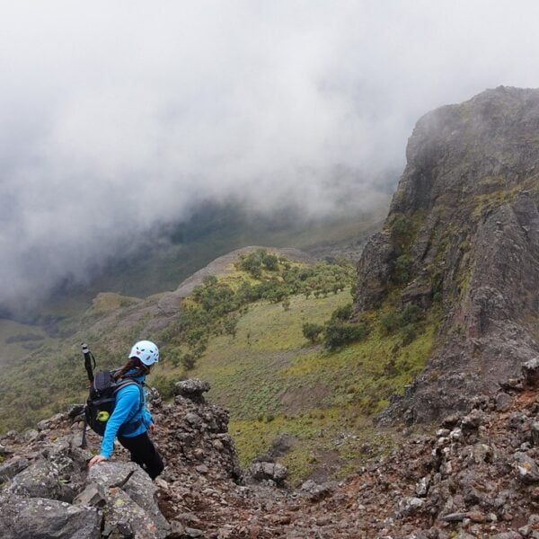 Two hikers with helmets and backpacks standing on a rocky mountain terrain, observing the misty landscape with a large rock formation nearby.