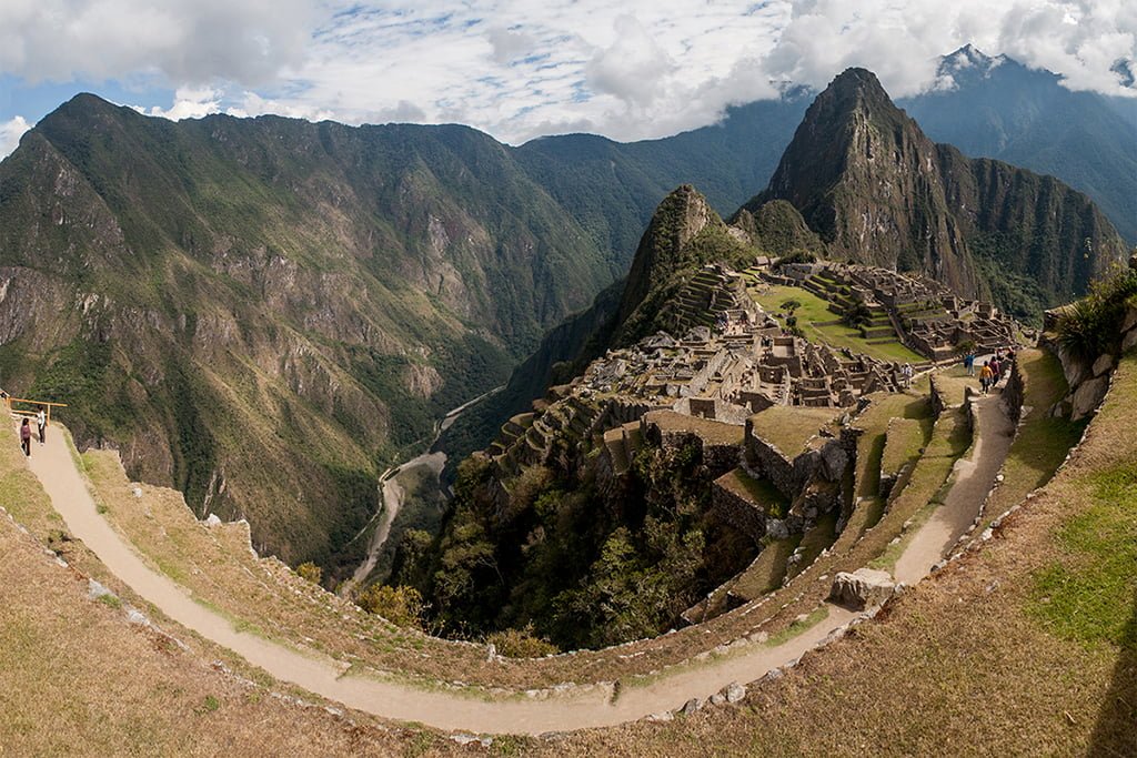 Panoramic view of Machu Picchu, showing ancient Incan ruins on terraced hillsides with tourists exploring, surrounded by the Andes Mountains under a partly cloudy sky.