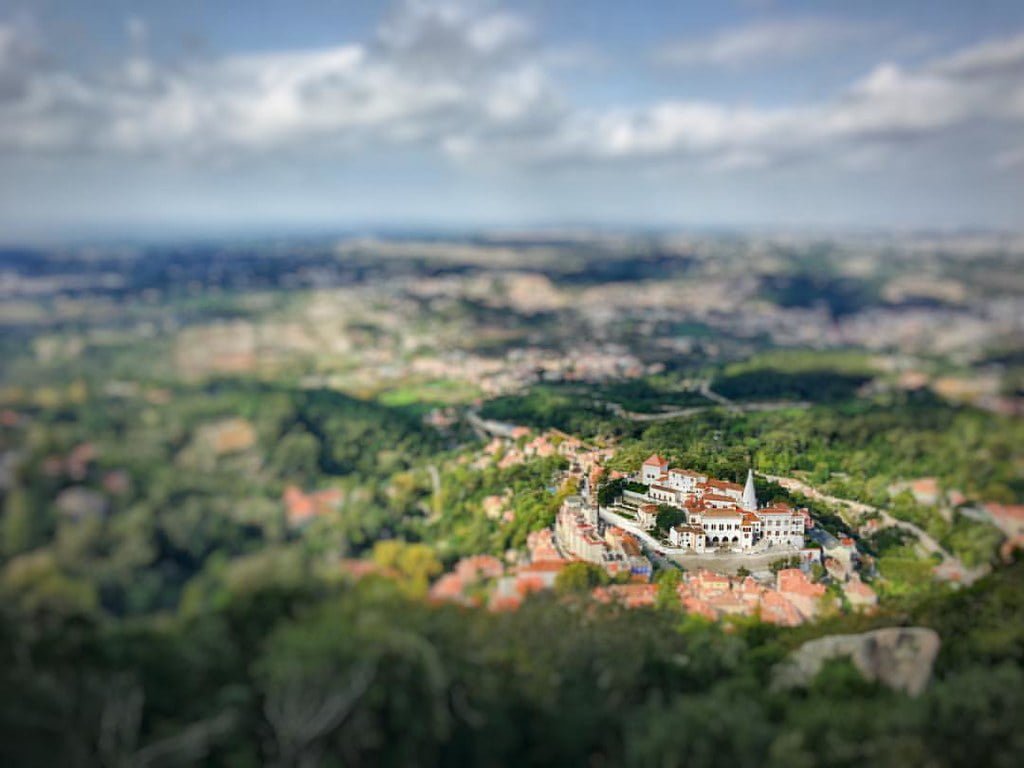 Aerial view of a white historical building complex with red roofs surrounded by greenery, using a tilt-shift photography effect that creates a miniature appearance.
