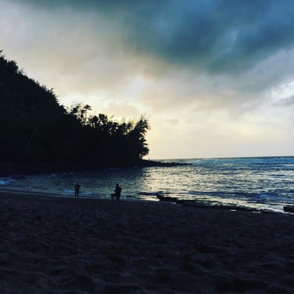 Alt text: A serene beach at dusk with silhouettes of people and trees against a backdrop of a cloudy sky and calm sea.