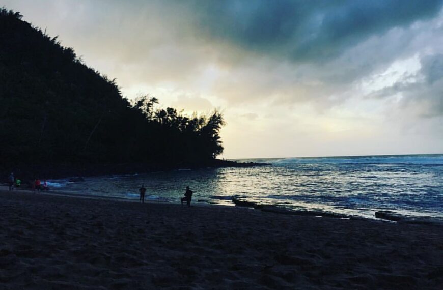 Alt text: A serene beach at dusk with silhouettes of people and trees against a backdrop of a cloudy sky and calm sea.
