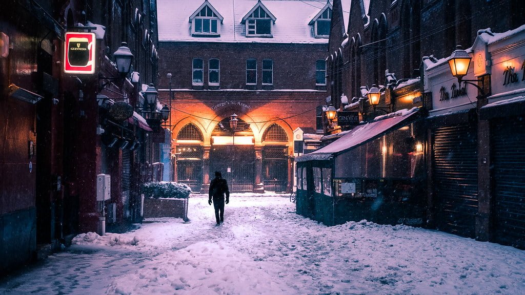 A person walking through a snow-covered street lined with closed shops and warm glowing street lamps at twilight, with a brick building featuring archways in the background.