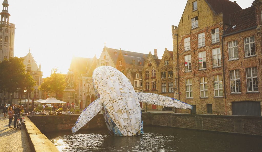 A large whale sculpture made of white and blue plastic waste appears to be jumping out of the canal in front of historical European buildings bathed in warm sunlight. People are visible enjoying the outdoor setting near the canal's edge.