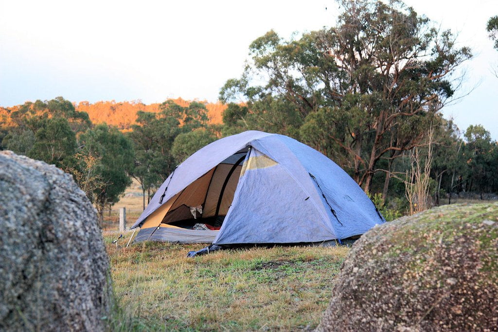 A blue camping tent pitched in a natural grassy area surrounded by trees and rocks, with a hilly background lit by the soft glow of sunrise or sunset.