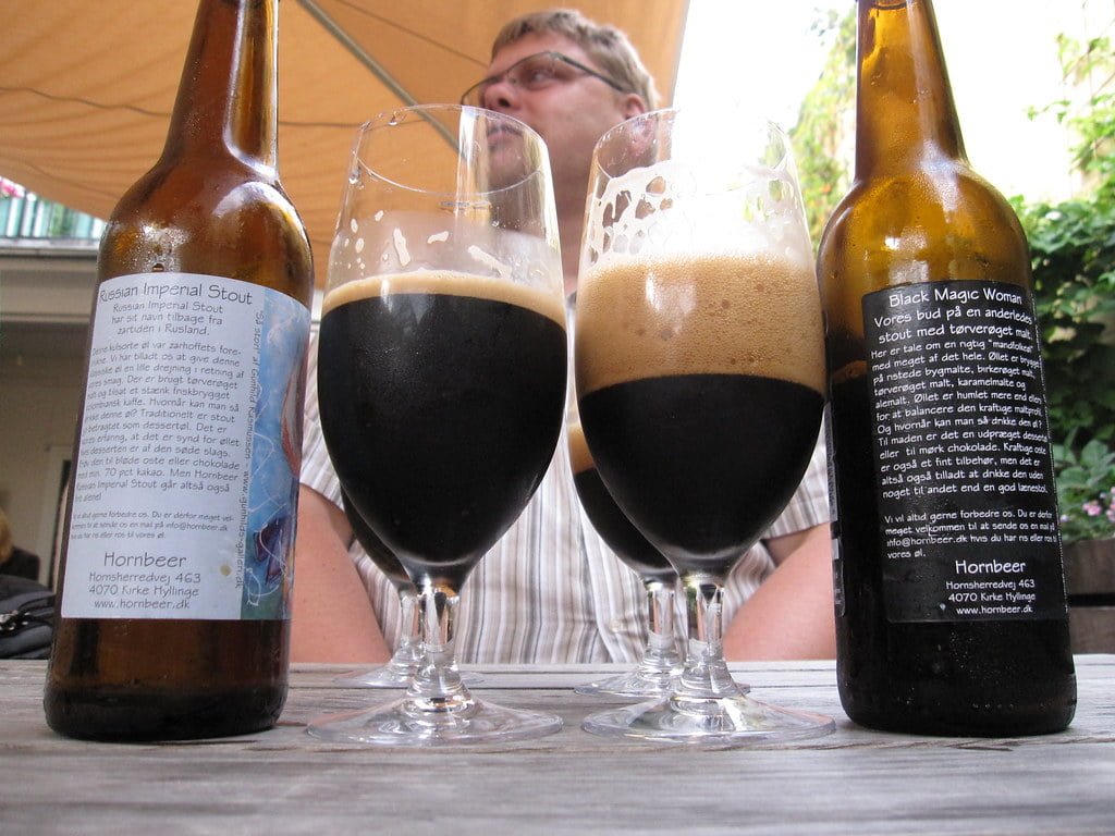 Two bottles of beer with labels "Russian Imperial Stout" and "Black Magic Woman" resting on a table alongside two glasses filled with dark beer, with a man in the background.