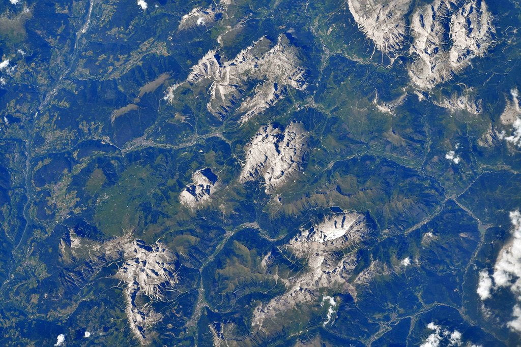 Satellite image showing rugged, snow-capped mountain ranges amidst green forested areas with some scattered clouds.