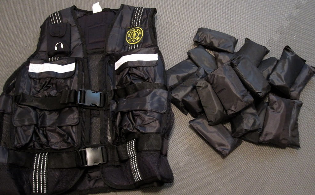 A black tactical vest with white reflective stripes and a badge on the front, next to a pile of separate pouches that appear to be weights, all laid out on a grey background.