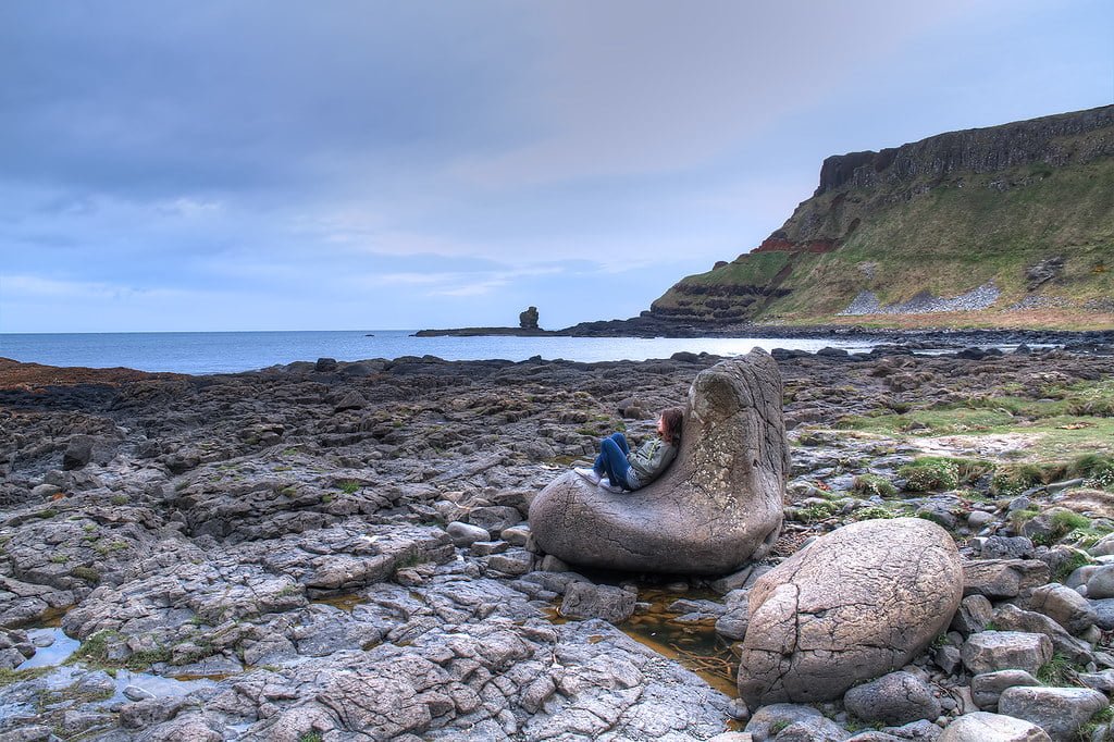 Person sitting in a natural rock formation resembling a chair along a rugged coast with cliffs, rocks, and the sea under a cloudy sky.