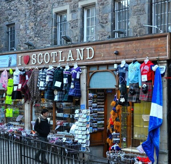 A storefront of "I Love Scotland" shop with various Scottish souvenirs on display, including clothes and accessories, next to another shop named "Castle." A person is standing outside the store.