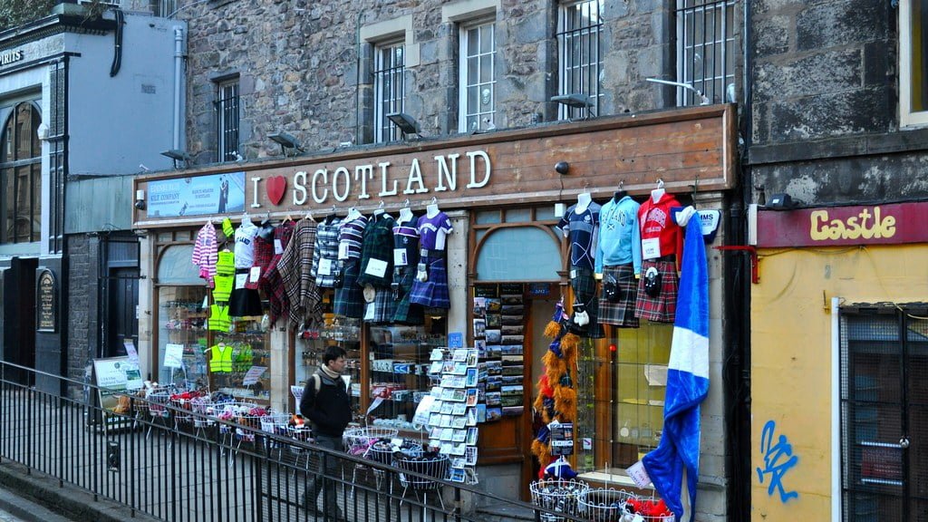 A storefront of "I Love Scotland" shop with various Scottish souvenirs on display, including clothes and accessories, next to another shop named "Castle." A person is standing outside the store.