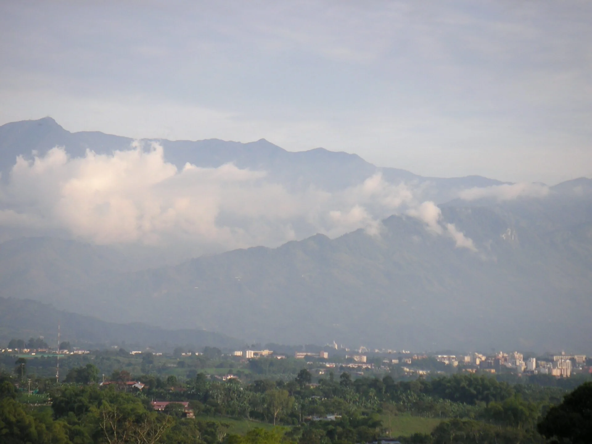A landscape view of a town with buildings spread out amid greenery, nestled at the base of a mountain range with clouds partially covering the peaks.