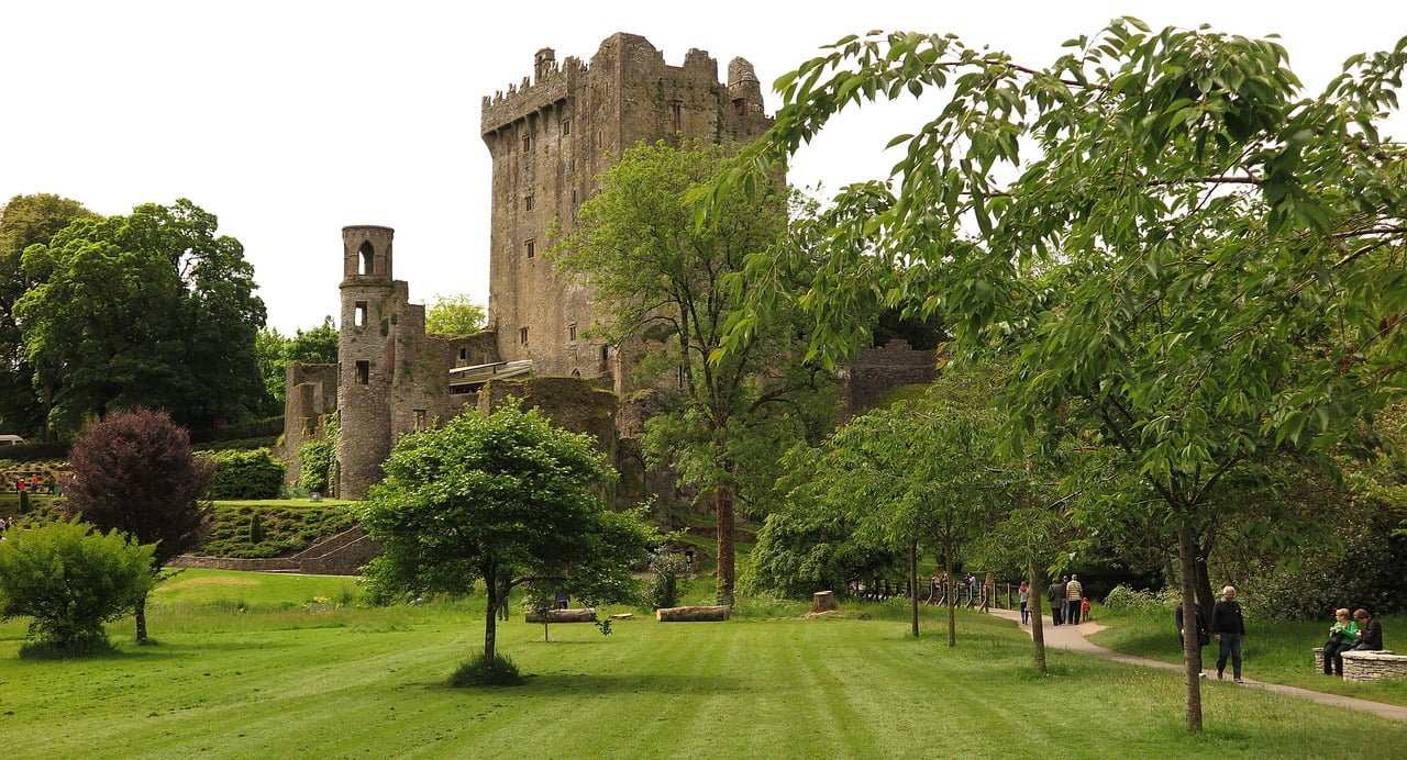 Alt text: A lush green park with visitors walking and sitting on benches, a well-maintained lawn, and trees in the foreground, with the ruins of an ancient towering stone castle in the background under a cloudy sky.