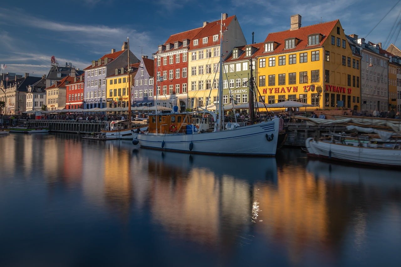 A scenic view of Nyhavn in Copenhagen, Denmark, with colorful buildings reflecting in the calm water and moored boats along the quayside.