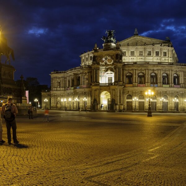 An illuminated historical building at night with an ornate street lamp in the foreground and a couple of people standing nearby.
