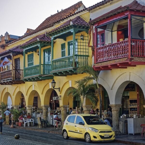 Street view of colorful colonial-style buildings with archways and balconies, people socializing and walking, and a yellow taxi parked in front.