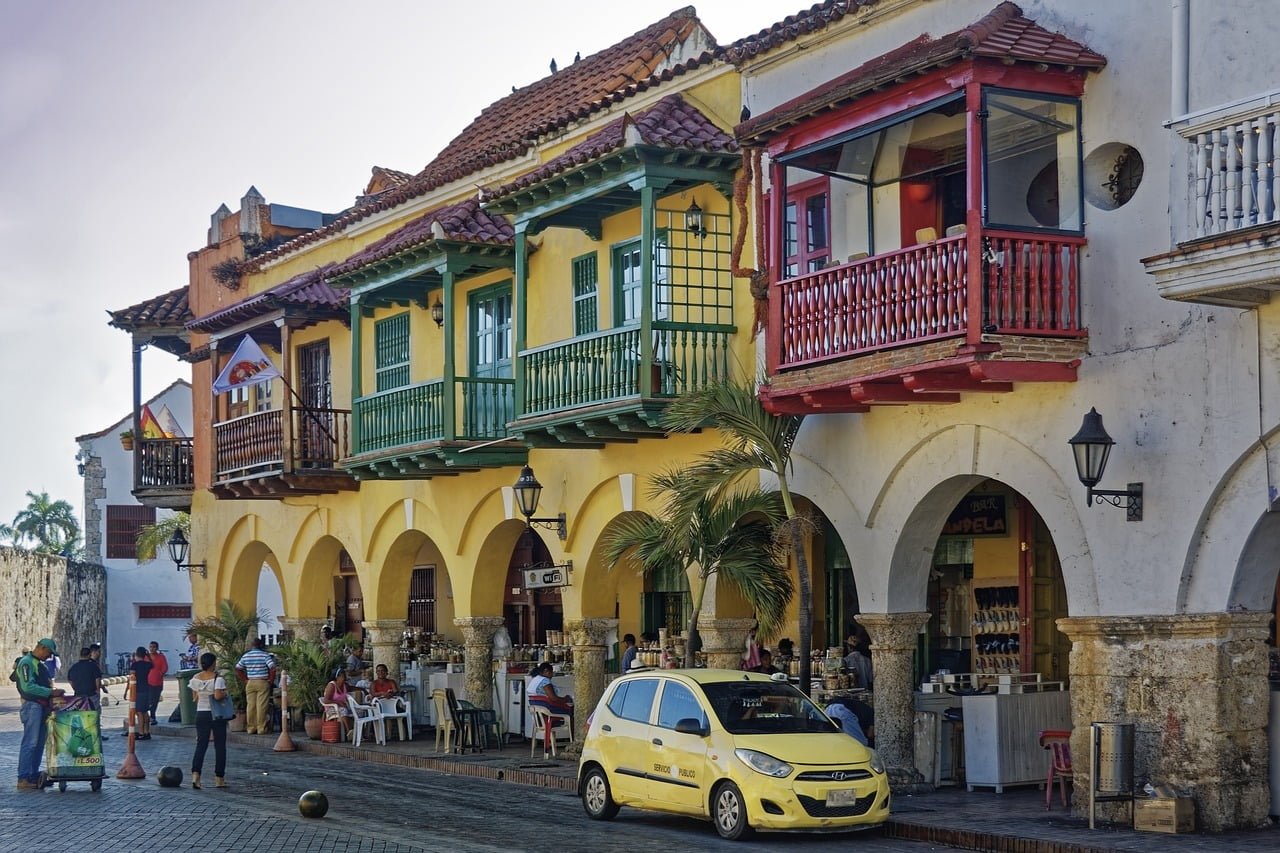 Street view of colorful colonial-style buildings with archways and balconies, people socializing and walking, and a yellow taxi parked in front.