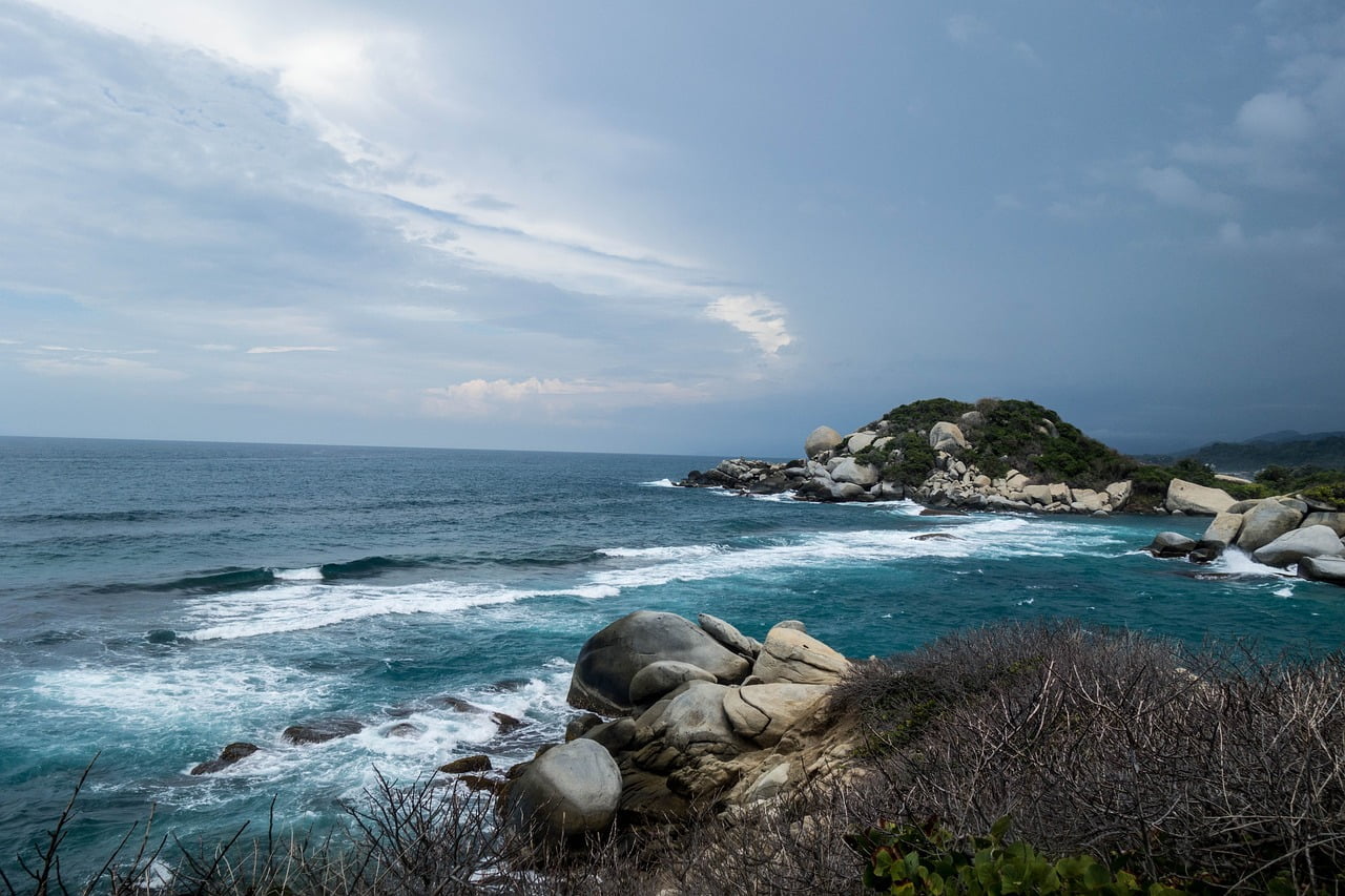 A coastal seascape with turbulent blue waters crashing against a rocky shore lined with large boulders under a cloudy sky.