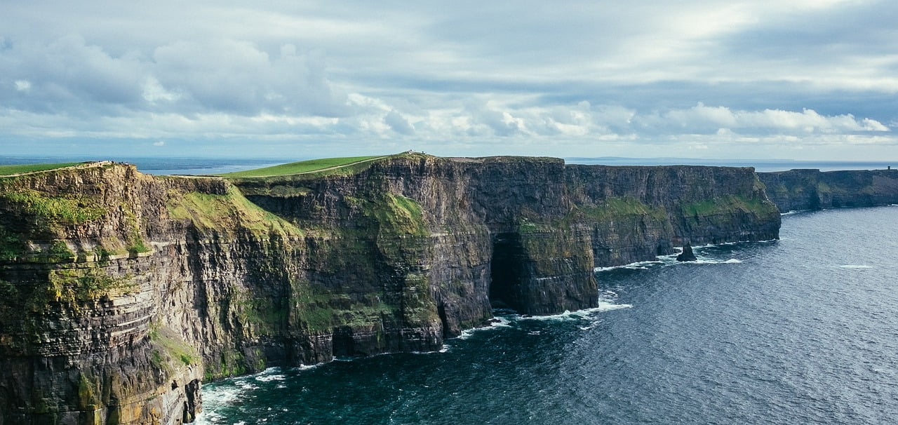 A panoramic view of the Cliffs of Moher under a cloudy sky, showcasing the lush green tops and rugged cliff faces overlooking the Atlantic Ocean.