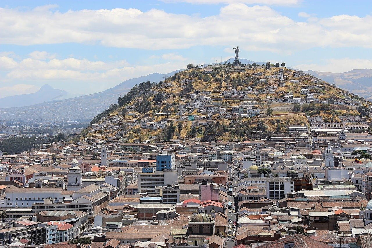 A panoramic view of a dense urban area with buildings covering the hill slopes, leading up to a statue on a hilltop under a clear sky.
