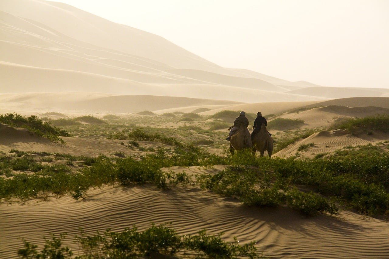 Two individuals riding camels across a desert landscape with sand dunes and sparse vegetation in the foreground, under a hazy sky.