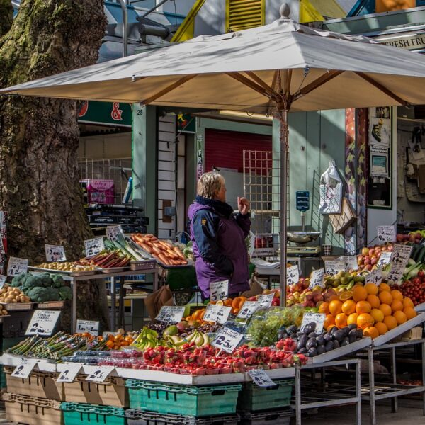 A bustling outdoor market with fresh fruit and vegetable stalls under white umbrellas, featuring a customer and a vendor amidst colorful produce displays with price tags.