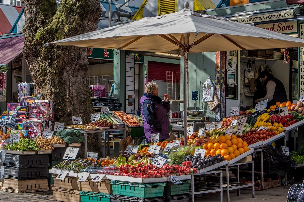 A bustling outdoor market with fresh fruit and vegetable stalls under white umbrellas, featuring a customer and a vendor amidst colorful produce displays with price tags.