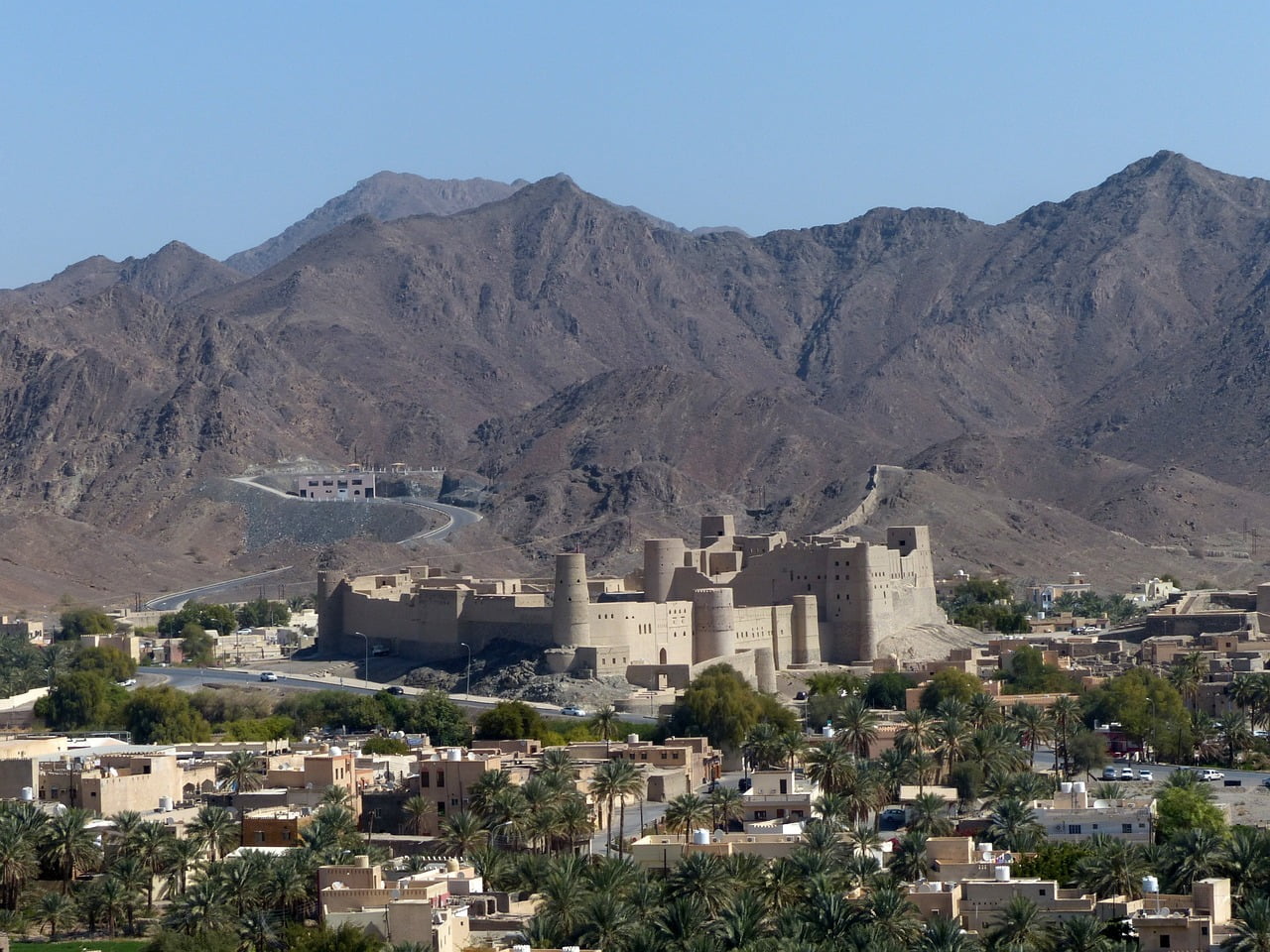 An ancient fortress stands prominently in front of a rugged mountain range, overlooking a town with low-rise buildings and palm trees.
