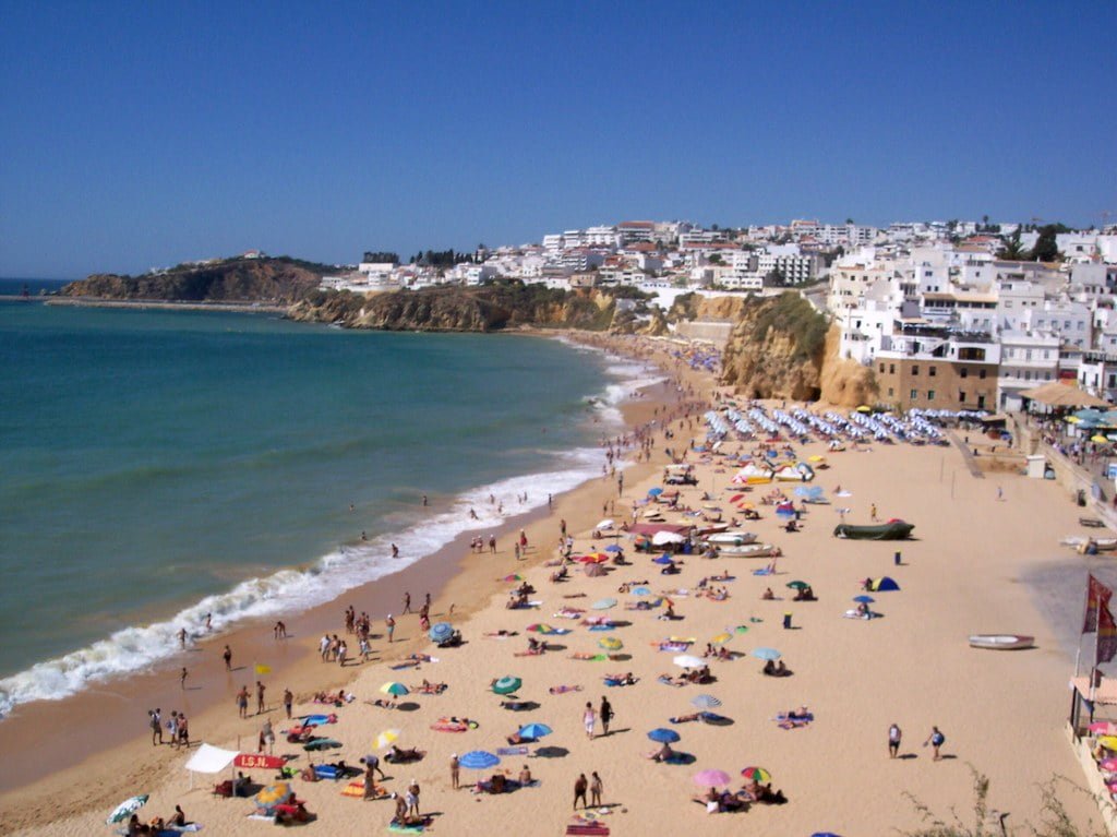 A bustling beach scene with numerous beachgoers and colorful umbrellas on the sand, adjacent to a coastal town with white buildings cascading down a hillside to the shoreline.