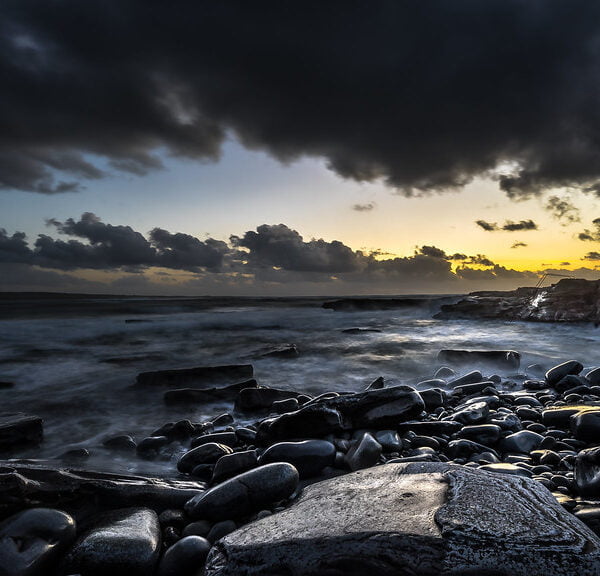 A dramatic seascape with a cloud-streaked sky at sunset, waves crashing onto a rocky shore lined with smooth pebbles and boulders, hint of sunlight piercing through to illuminate the horizon.