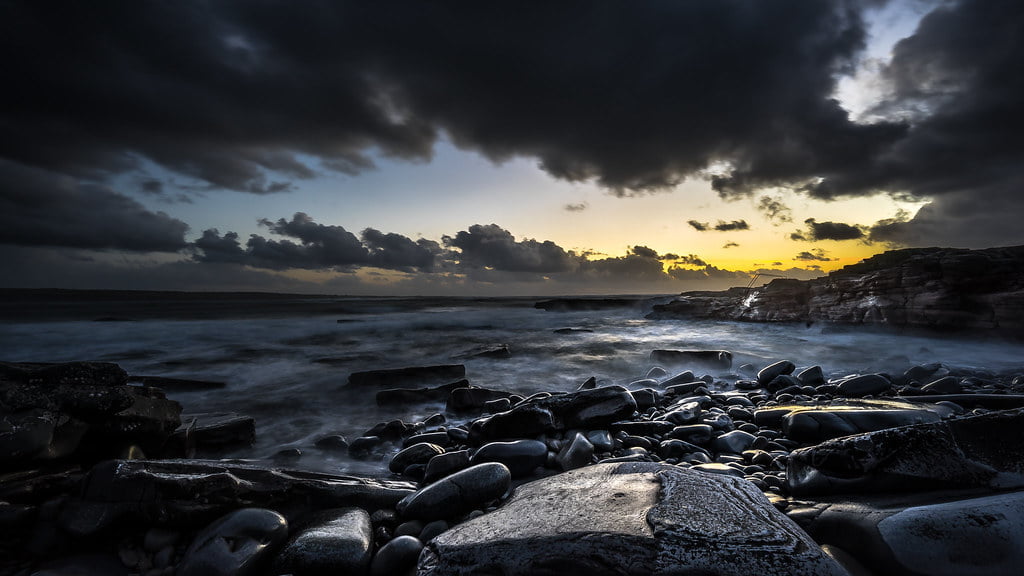 A dramatic seascape with a cloud-streaked sky at sunset, waves crashing onto a rocky shore lined with smooth pebbles and boulders, hint of sunlight piercing through to illuminate the horizon.