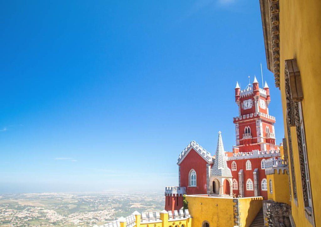 A view of the Pena Palace in Sintra, Portugal, showcasing its vibrant red and yellow walls under a clear blue sky.