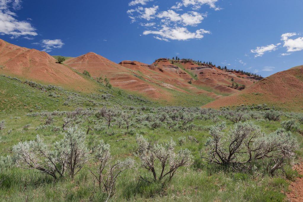 Red hills with sparse greenery under a blue sky with scattered clouds.