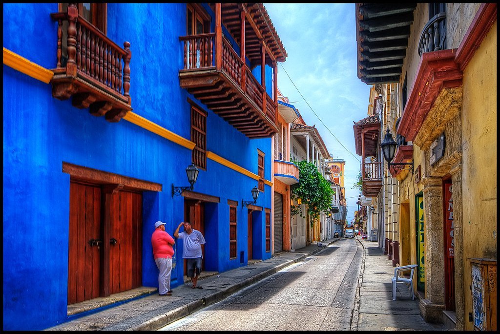 A vibrant street scene with cobblestone road and brightly painted colonial buildings, featuring a prominent blue wall with wooden balconies and doors, and two people standing by the doorway on the left.