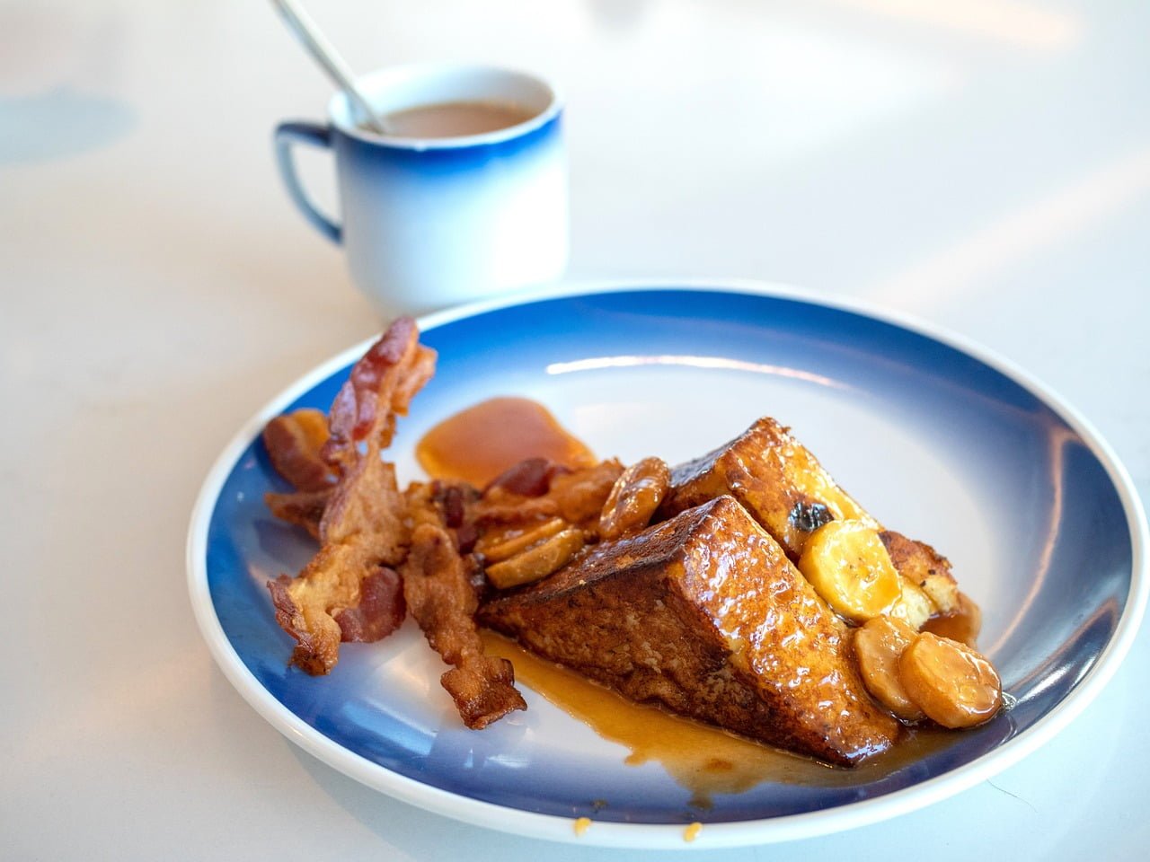 A plate of French toast topped with sliced bananas and syrup, accompanied by strips of bacon, with a cup of tea in the background.