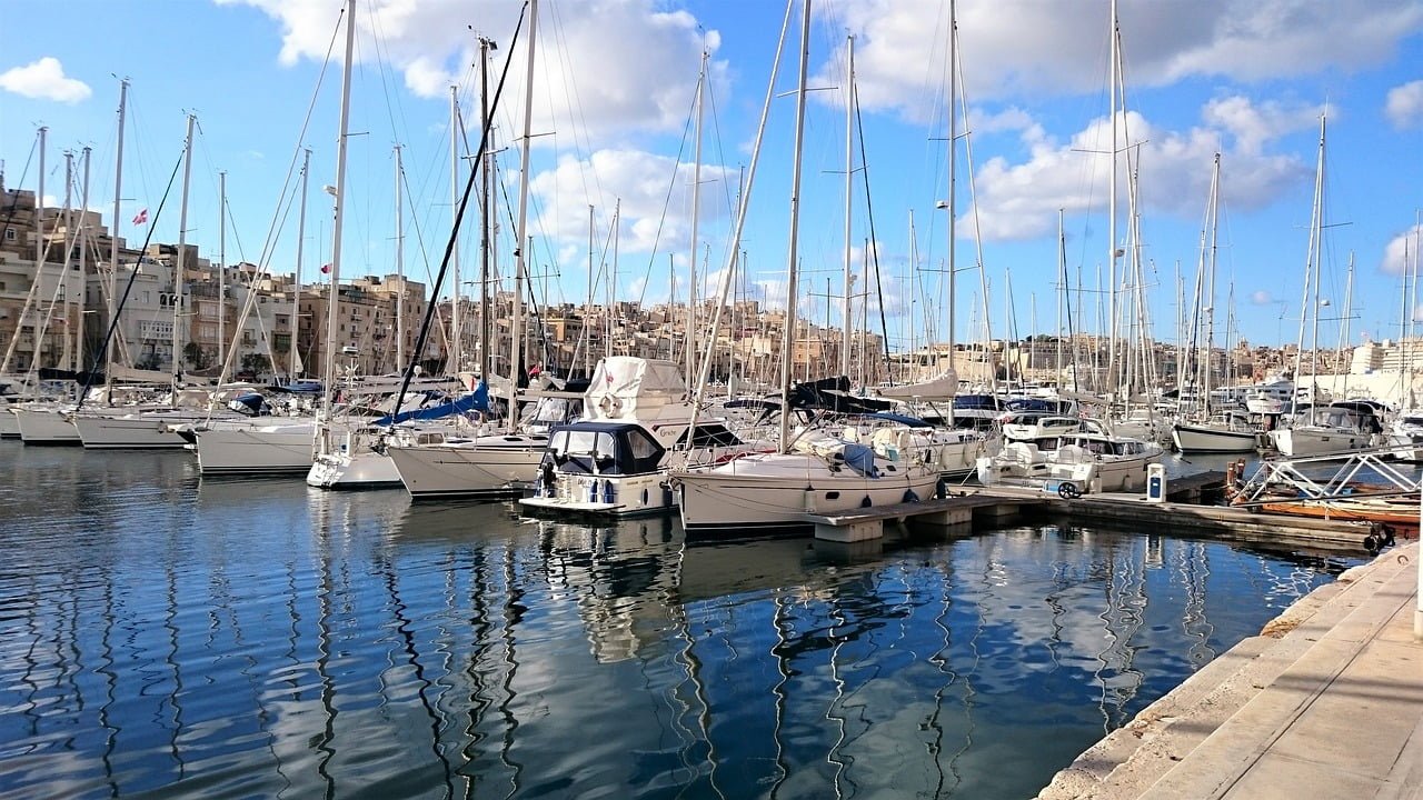 A marina filled with various sailing boats and yachts, with calm water reflecting the vessels and a cityscape in the background under a blue sky with scattered clouds.