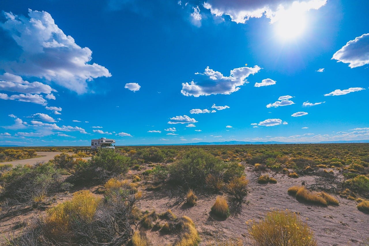 A sunny desert landscape with scattered clouds in the blue sky, and an RV parked beside a road in the distance.