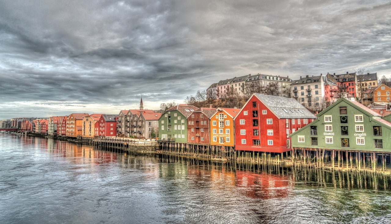 The image shows a row of colorful wooden houses on stilts along the waterfront, with a cloudy sky overhead reflecting in the calm water.