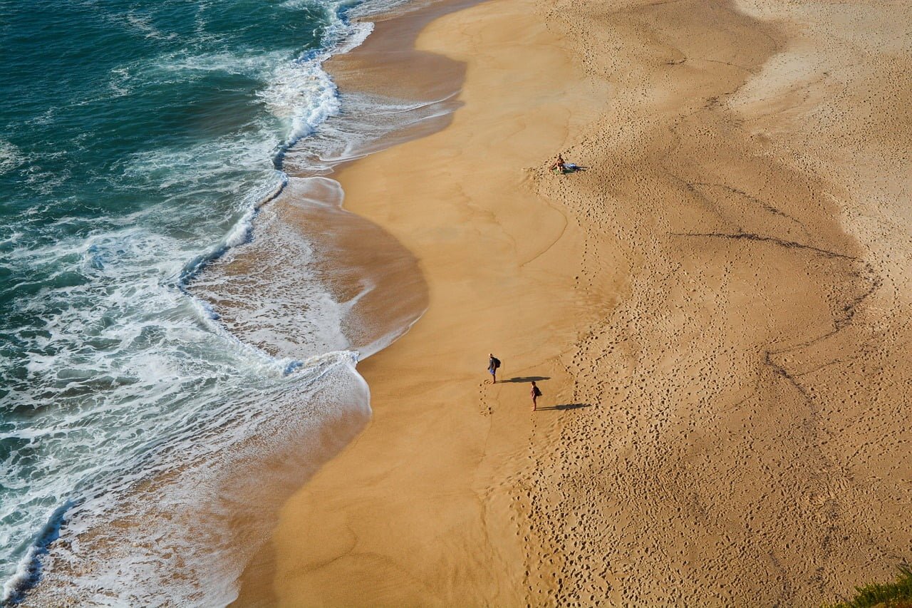 Aerial view of a sandy beach with waves lapping at the shore, two people walking, and a third person lying on a towel.