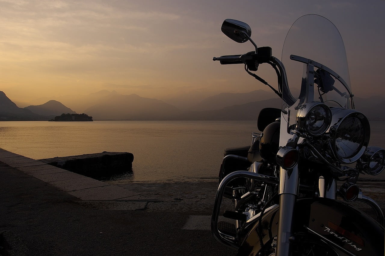 Alt text: A motorcycle parked on a lakeside at dusk, with mountains silhouetted in the background under a soft gradient sky from orange to purple.