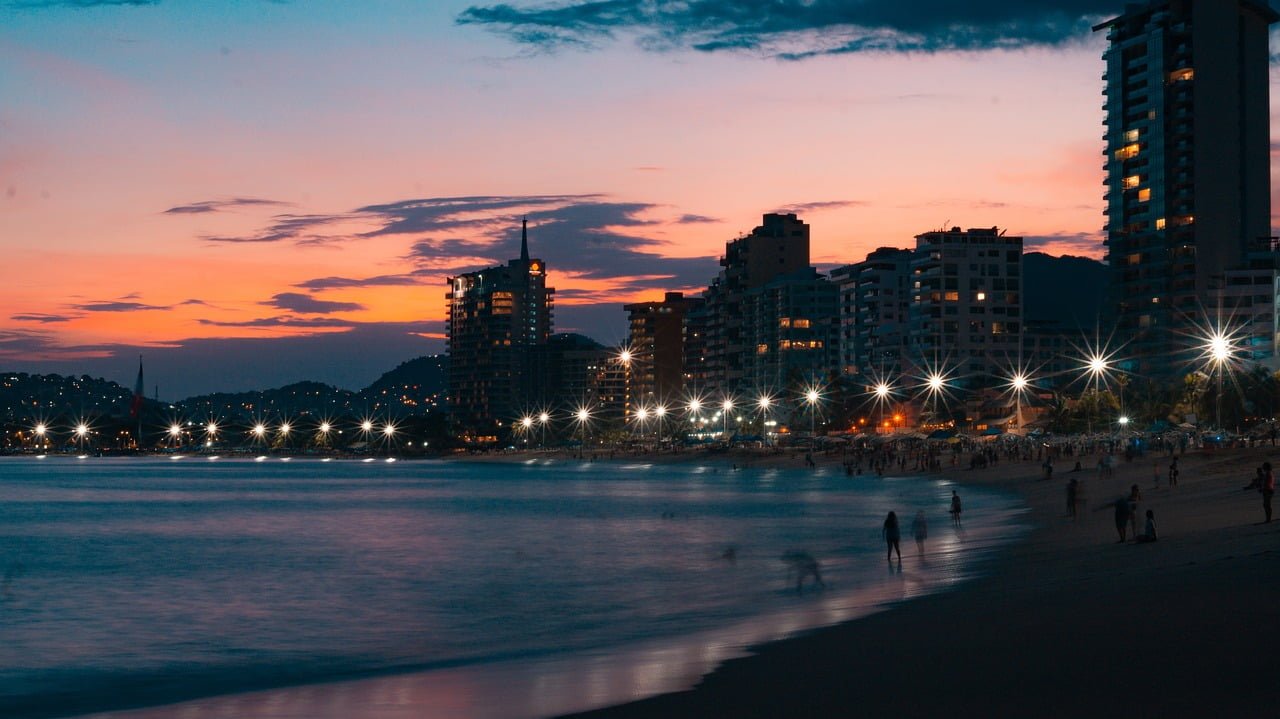 A beachside cityscape at twilight with colorful clouds in the sky, building lights twinkling, and silhouettes of people on the beach.