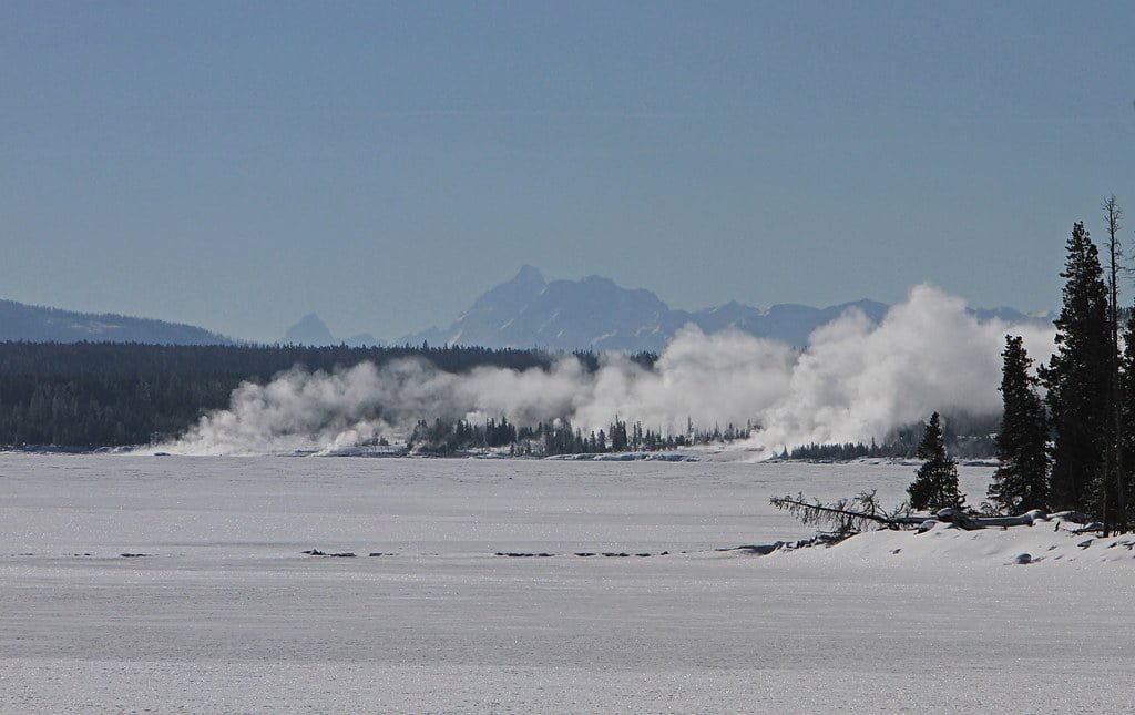 A snowy landscape with steaming geothermal features in the distance, set against a backdrop of mountain peaks and a clear blue sky.