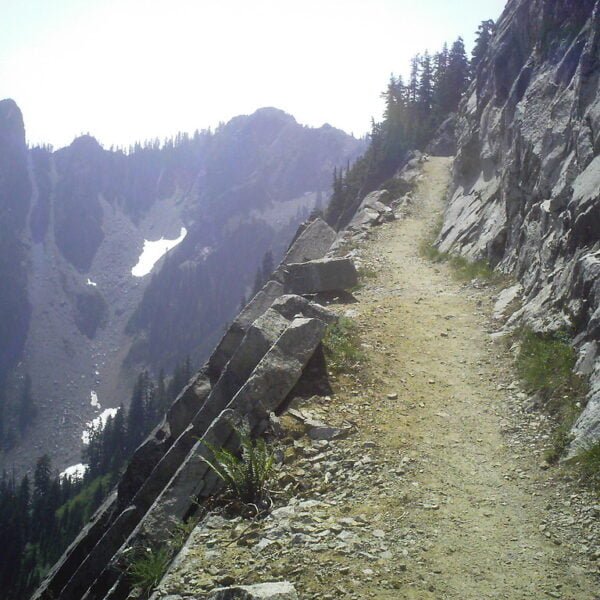 A mountain trail with a rocky slope on the right and a steep drop-off on the left, leading towards alpine terrain with patches of snow in the distance.