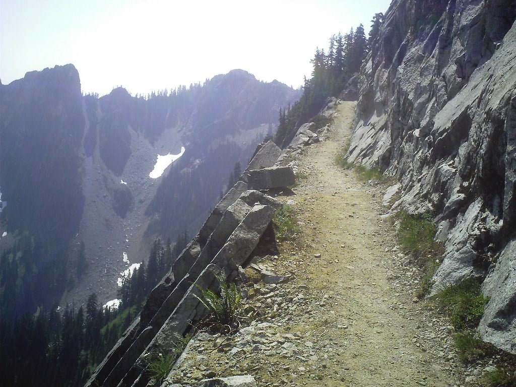 A mountain trail with a rocky slope on the right and a steep drop-off on the left, leading towards alpine terrain with patches of snow in the distance.