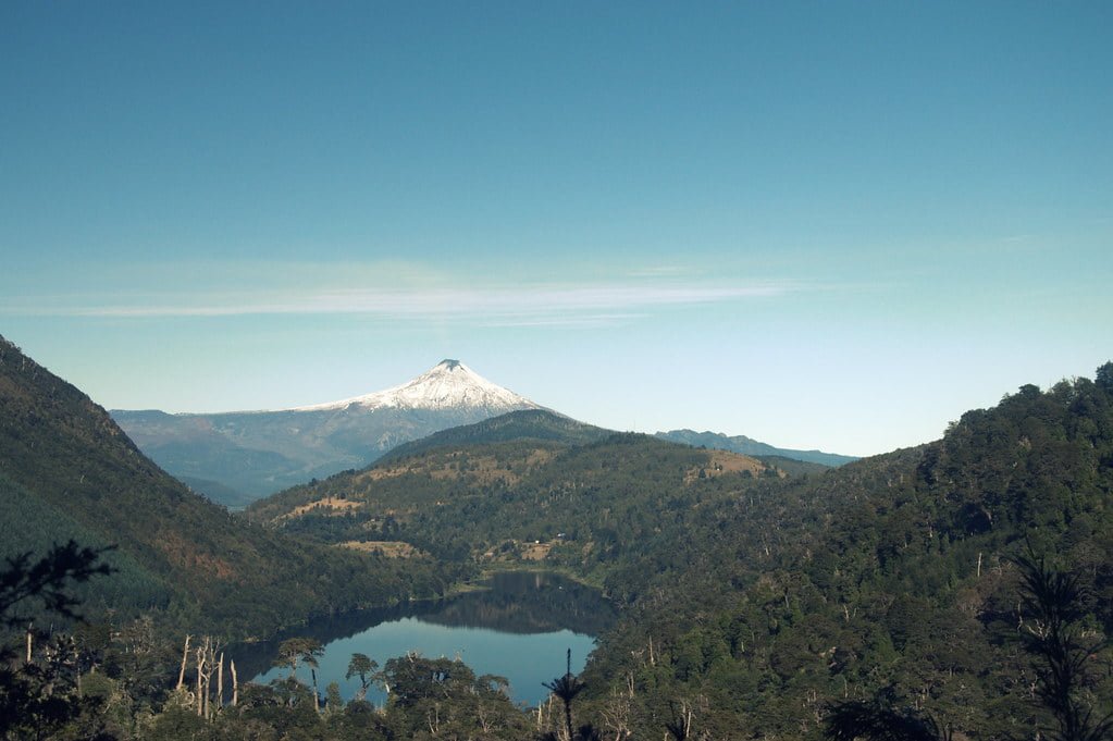 Alt text: A serene landscape featuring a snow-capped mountain in the background with a foreground of a forest and a reflective lake nestled between hills under a clear blue sky.