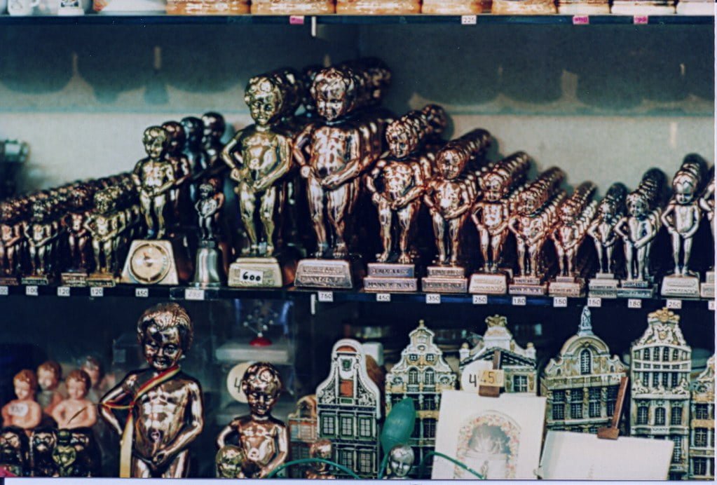 Rows of shiny miniature Manneken Pis statues displayed in various sizes on a store shelf with price tags, alongside other souvenir items including miniature buildings.
